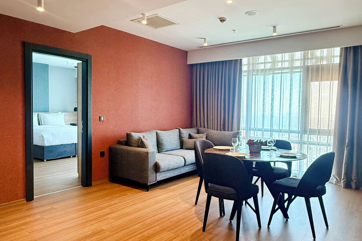 Ein modernes Hotelzimmer mit Holzboden, einer Couch, einem Esstisch mit Stühlen und einem großen Fenster mit Vorhängen.