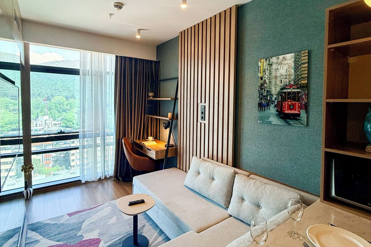Ein modernes Wohnzimmer mit einer Couch, einem Esstisch für zwei Personen, einem Sessel und Blick auf eine Stadtlandschaft durch große Fenster.