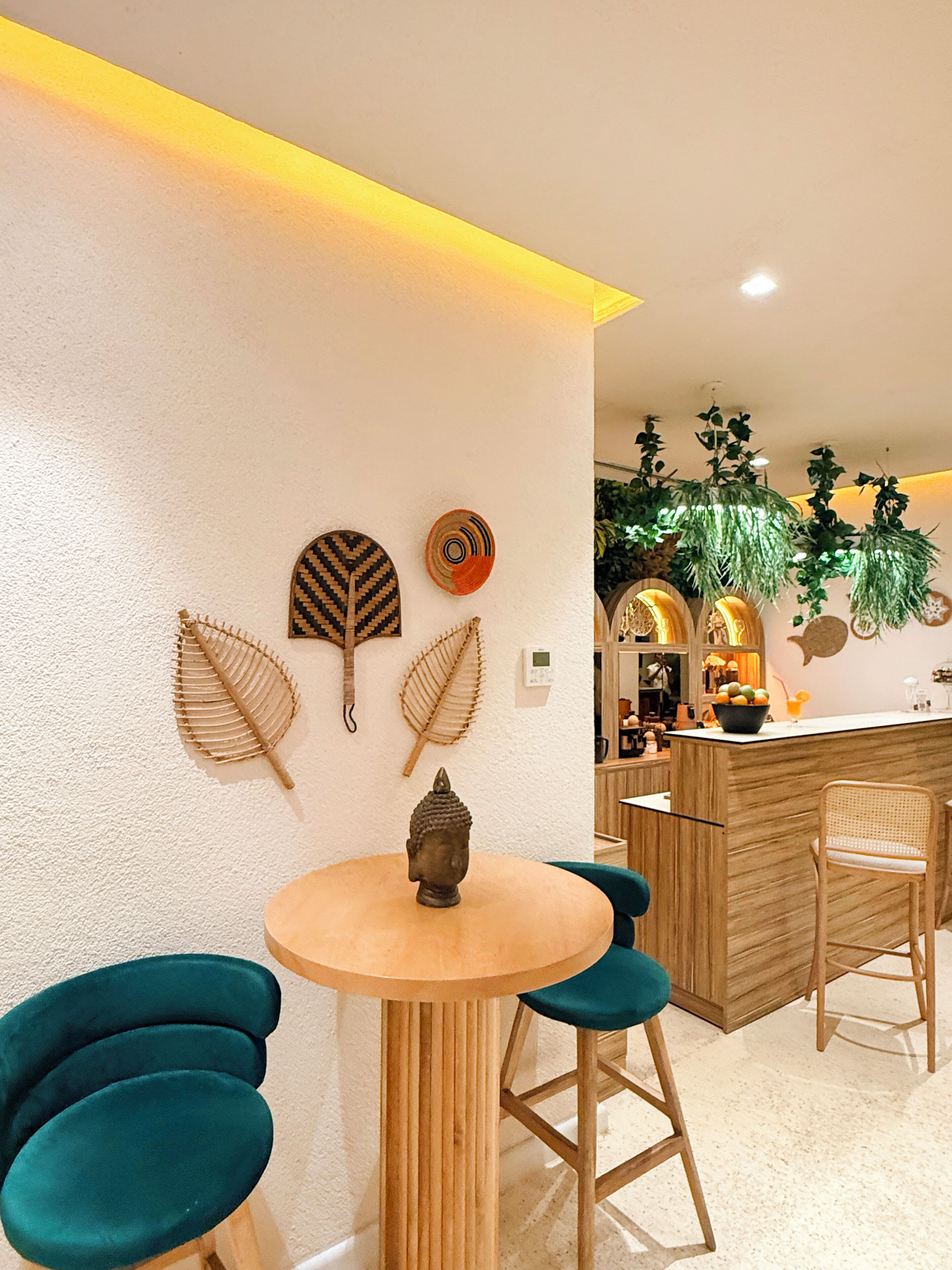 A modern interior with a round wooden table, teal chairs, decorative wall art, a wooden bar area, and hanging plants.