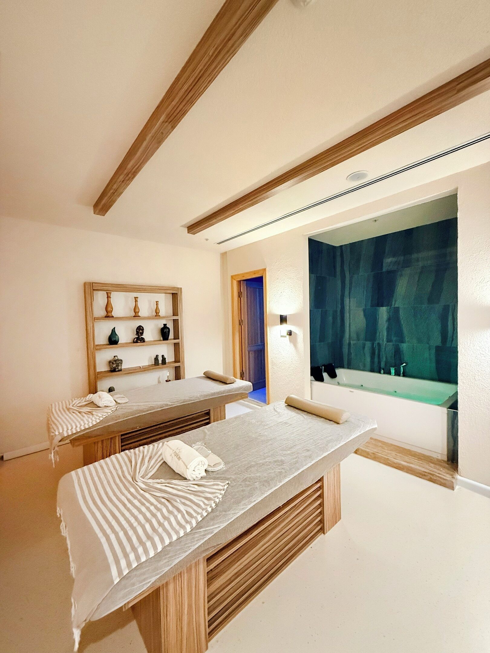 Un cuarto de spa con dos camillas de masaje, un estante de madera con adornos, un espejo grande y una bañera.