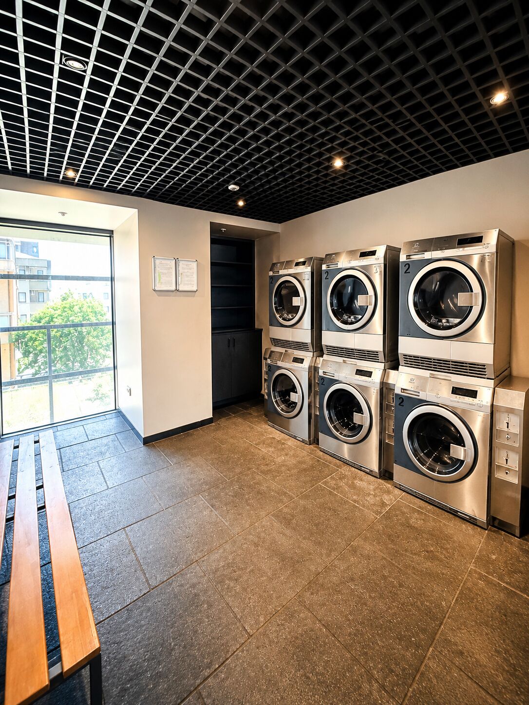 A laundry room with four stacked front-loading washers and dryers, a bench, and a view through a large window.