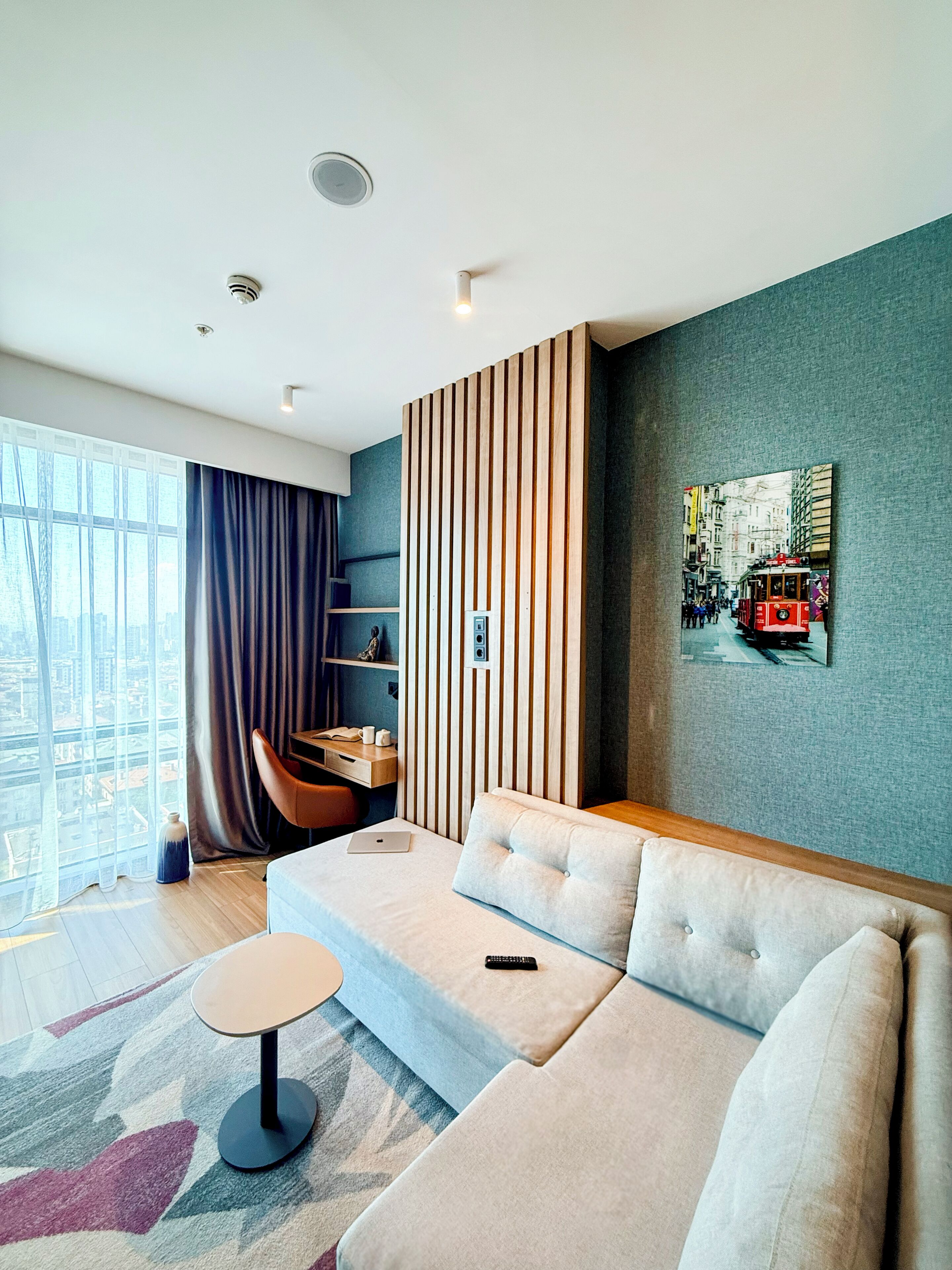 Una habitación de hotel moderna con sofá, una mesita redonda, un escritorio con silla, un ventanal con cortinas y vistas a la ciudad.