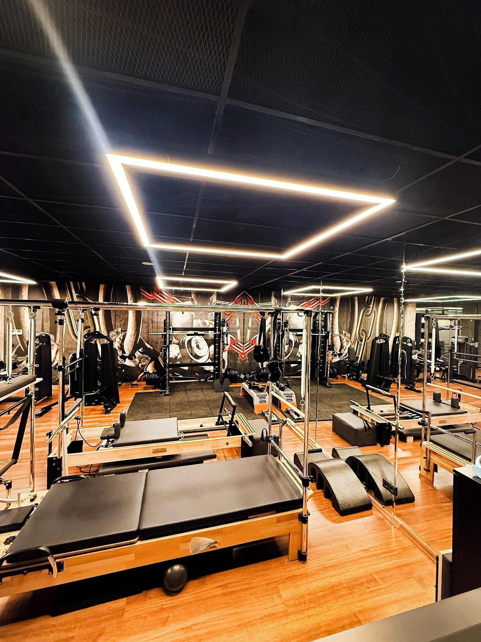 Ein Fitnessraum mit unterschiedlichen Trainingsgeräten, Holzboden und geometrischen Deckenleuchten.