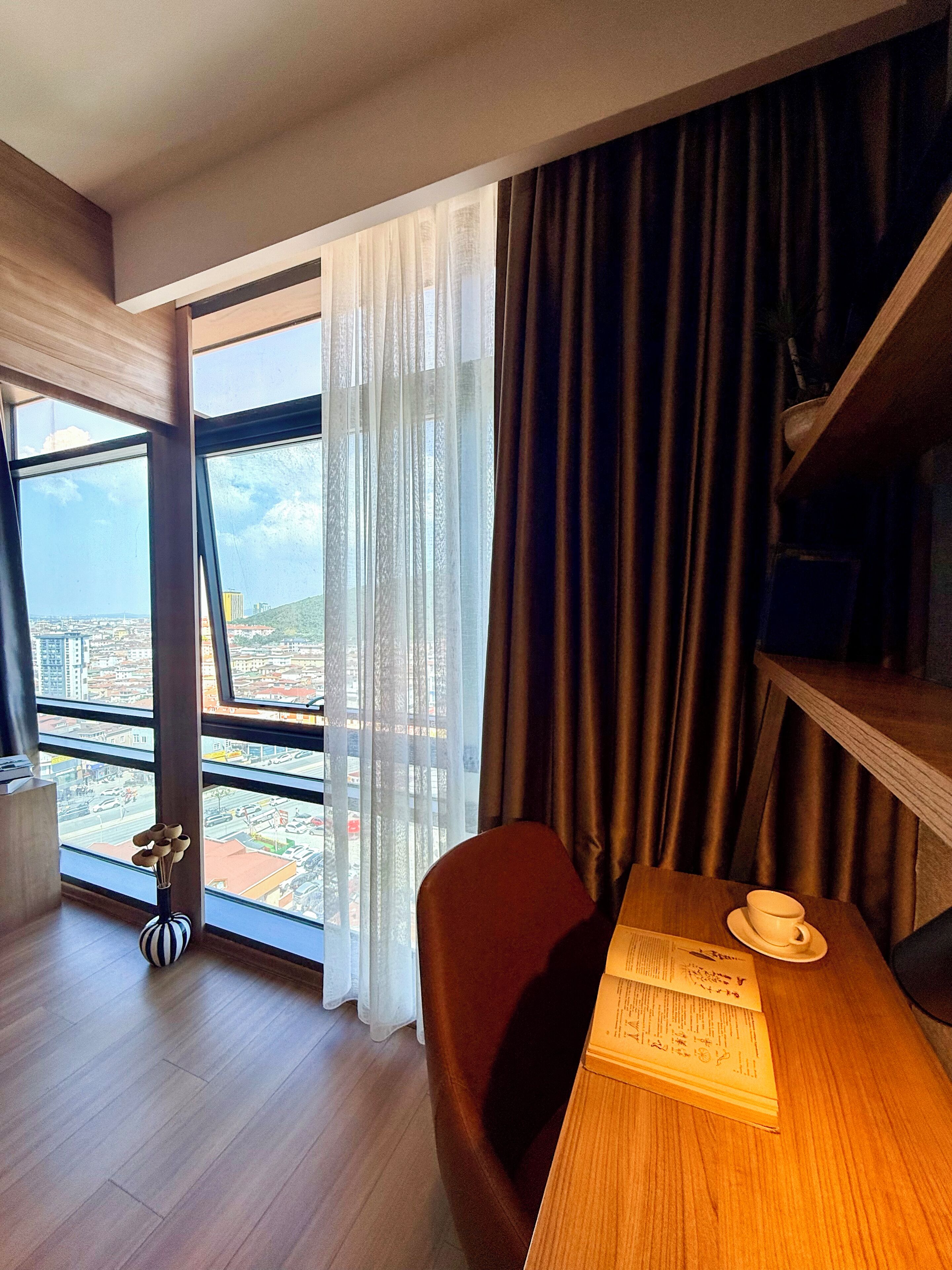 A hotel room with a large window, a wooden desk, a chair, and a vase with a plant.