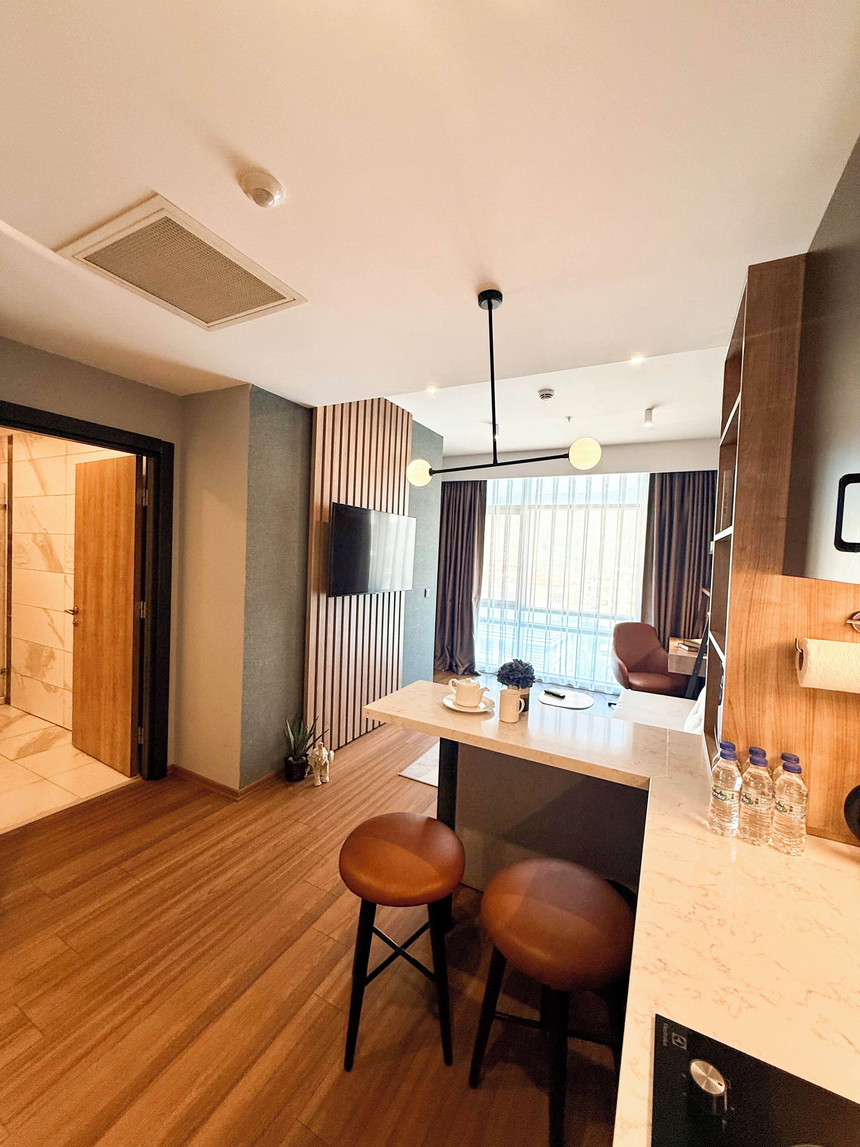 A modern hotel room with a wooden floor, a kitchenette, a seating area with stools, a TV, and a window with curtains.