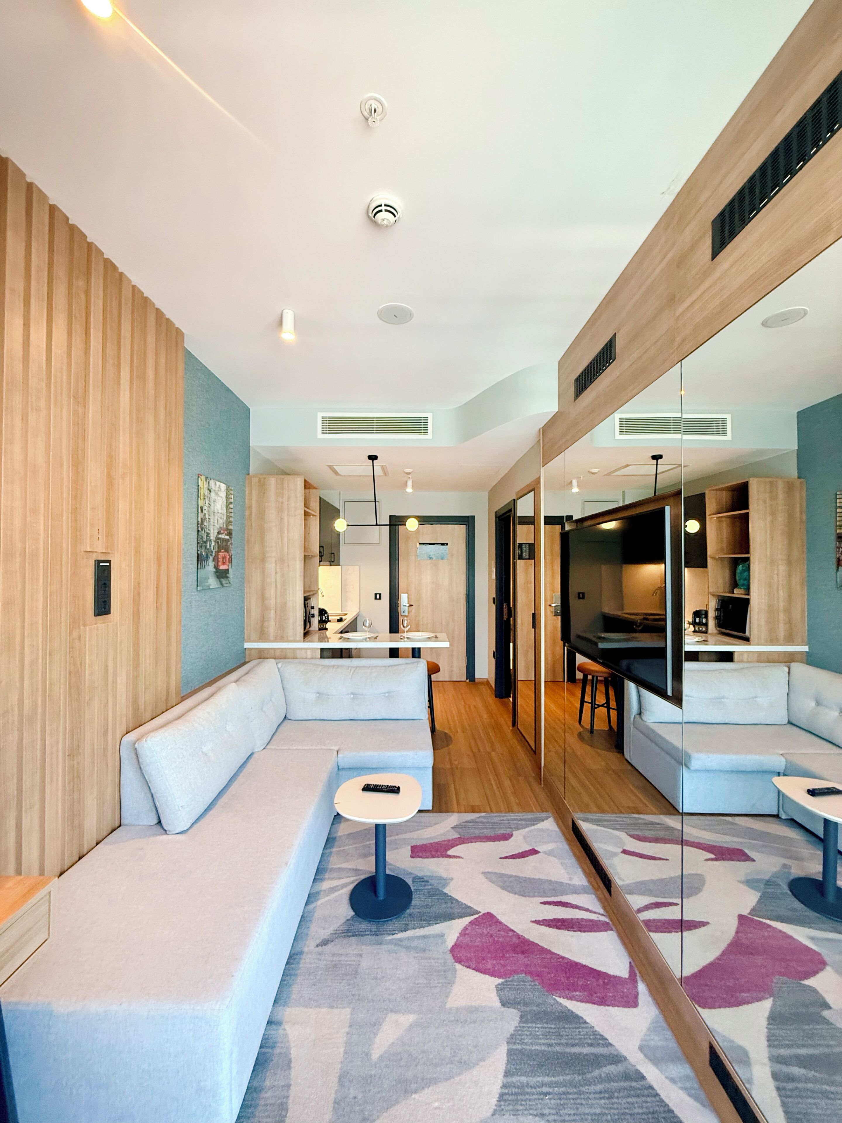 A modern living room with a sectional sofa, a small round table, a large mirror, and a kitchen area in the background.