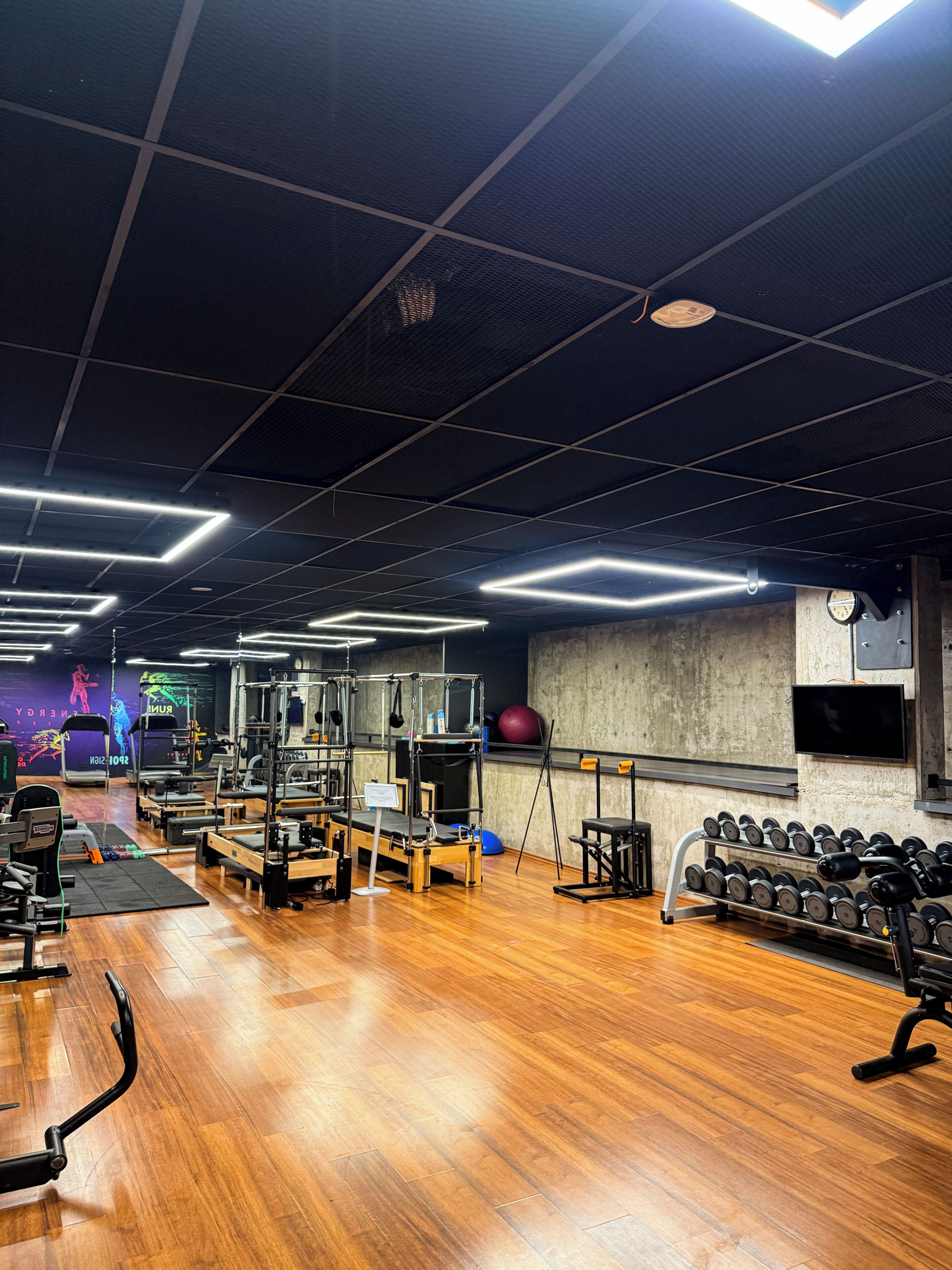 A gym with various exercise equipment, wooden flooring, and a ceiling with grid-like lighting fixtures.