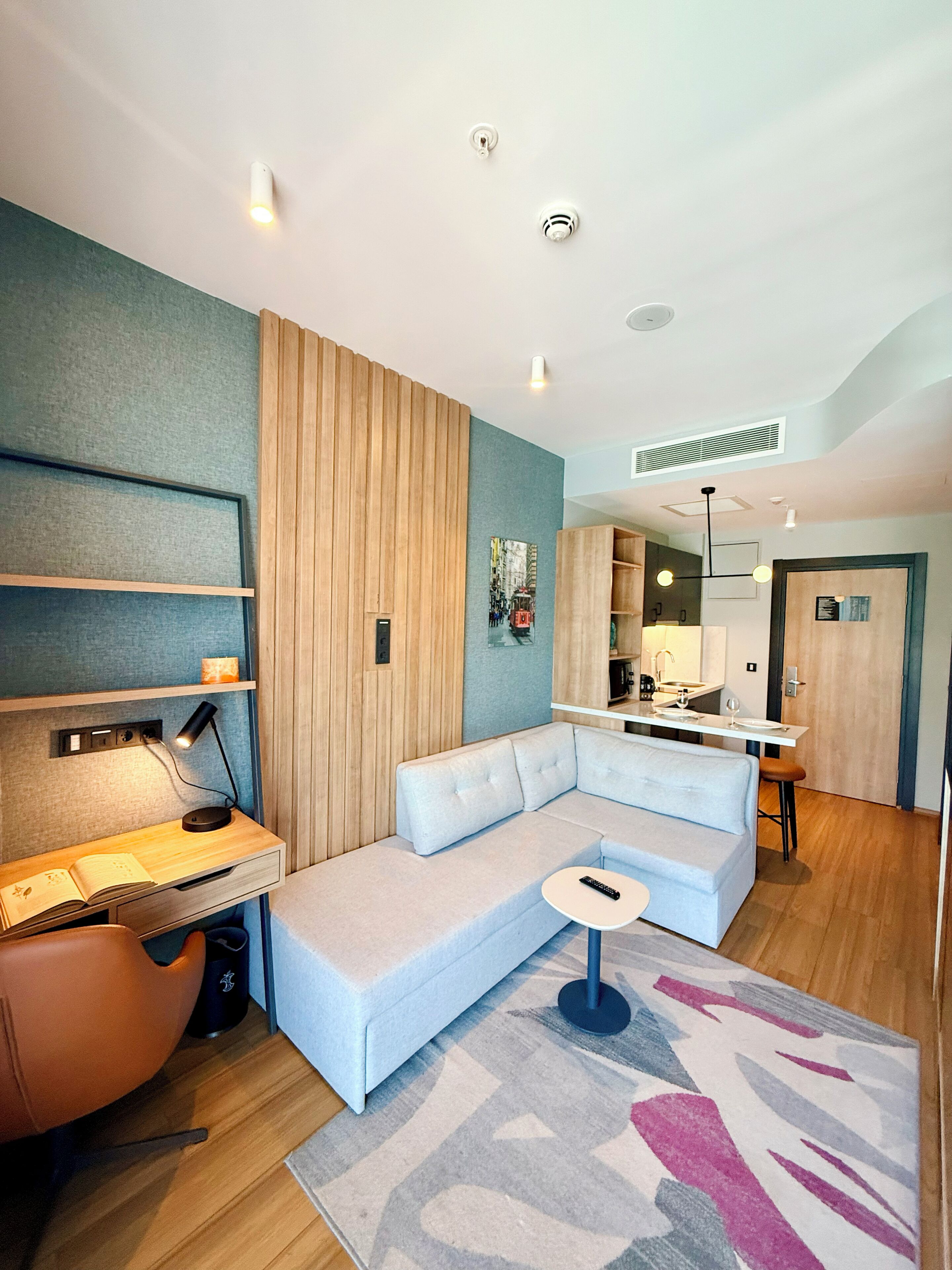 A modern living room with a sectional sofa, a desk with a lamp, a small round table, and a kitchen area in the background.