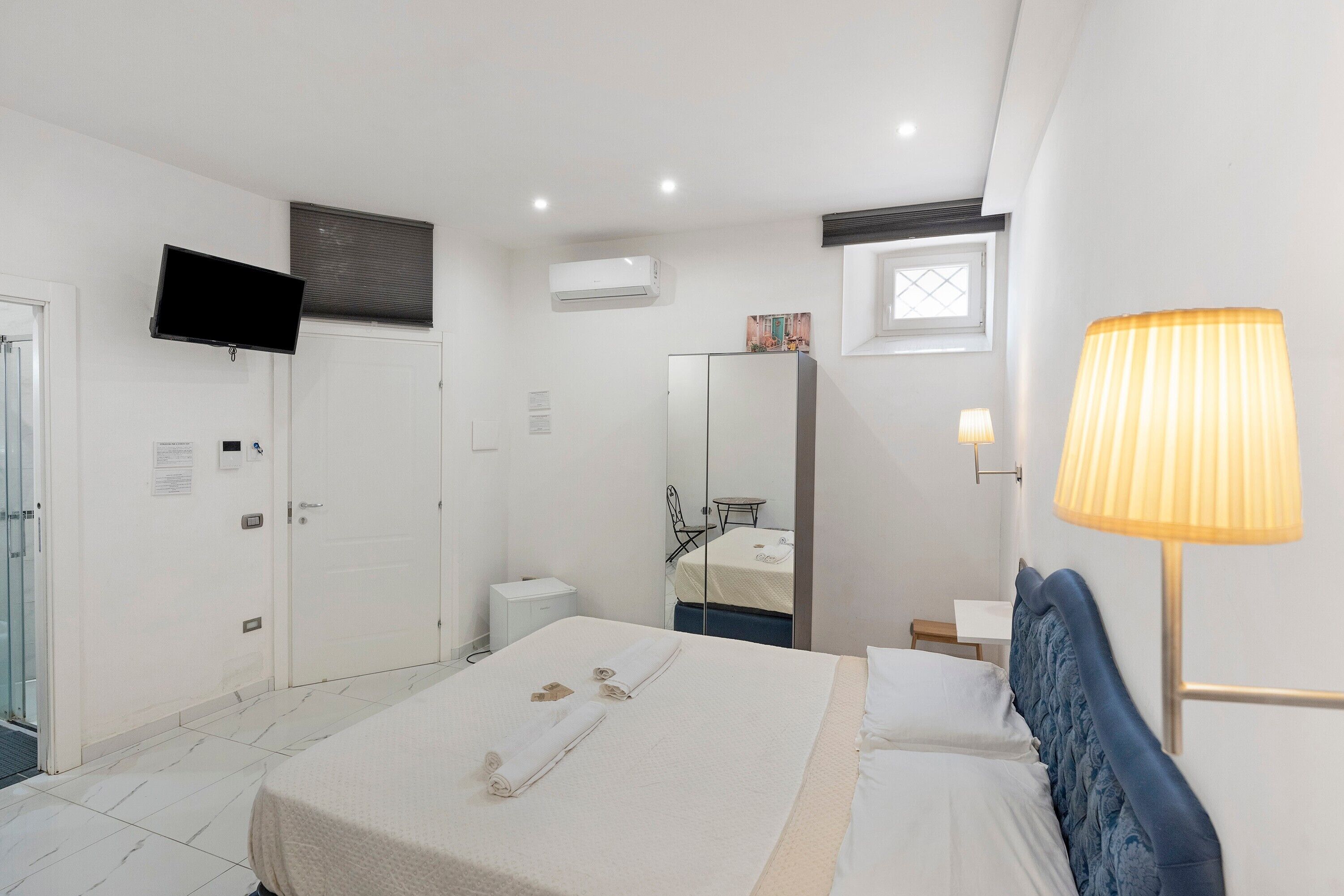 A hotel room with a bed, a bedside lamp, a television, an air conditioning unit, a small table, and a mirror.