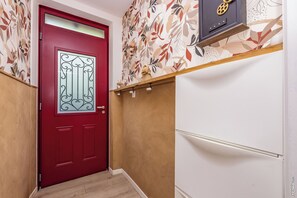 Eine rote Tür mit dekorativer Glasscheibe, eine Wand mit floralem Wandpapier und ein weißes Schränkchen mit einer Ablage darüber.