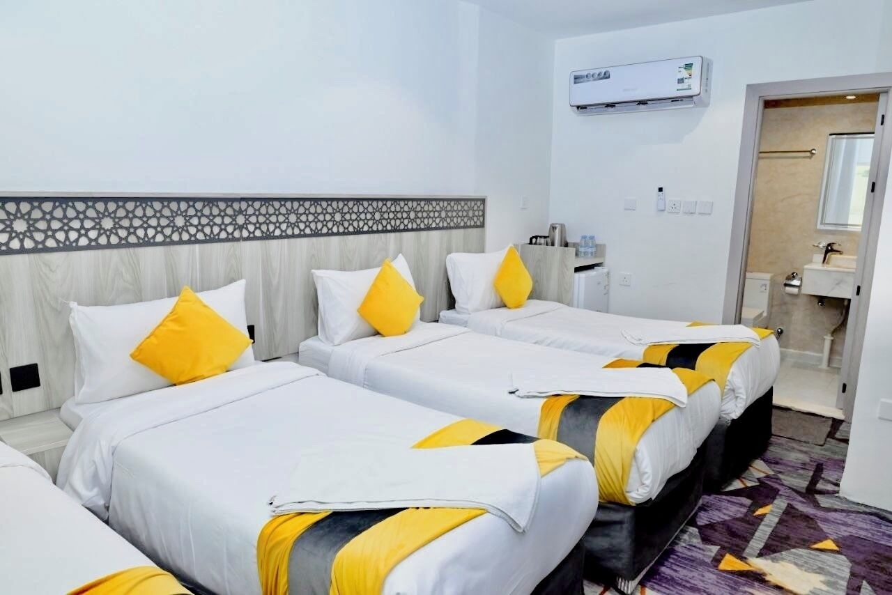 Habitació d'hotel amb tres llits, un panell decoratiu a la paret, un aparell d'aire condicionat i un bany visible a través d'una porta oberta.