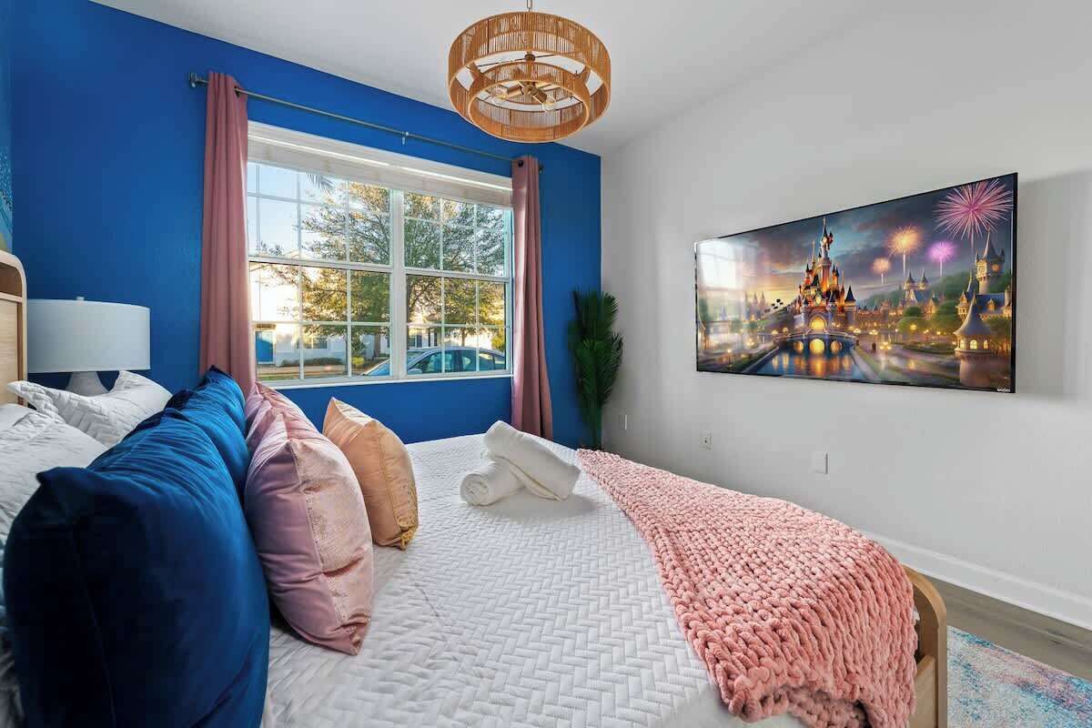 Un dormitorio con cama, almohadas, una ventana con cortinas, un televisor montado en la pared y un cuadro de fuegos artificiales.