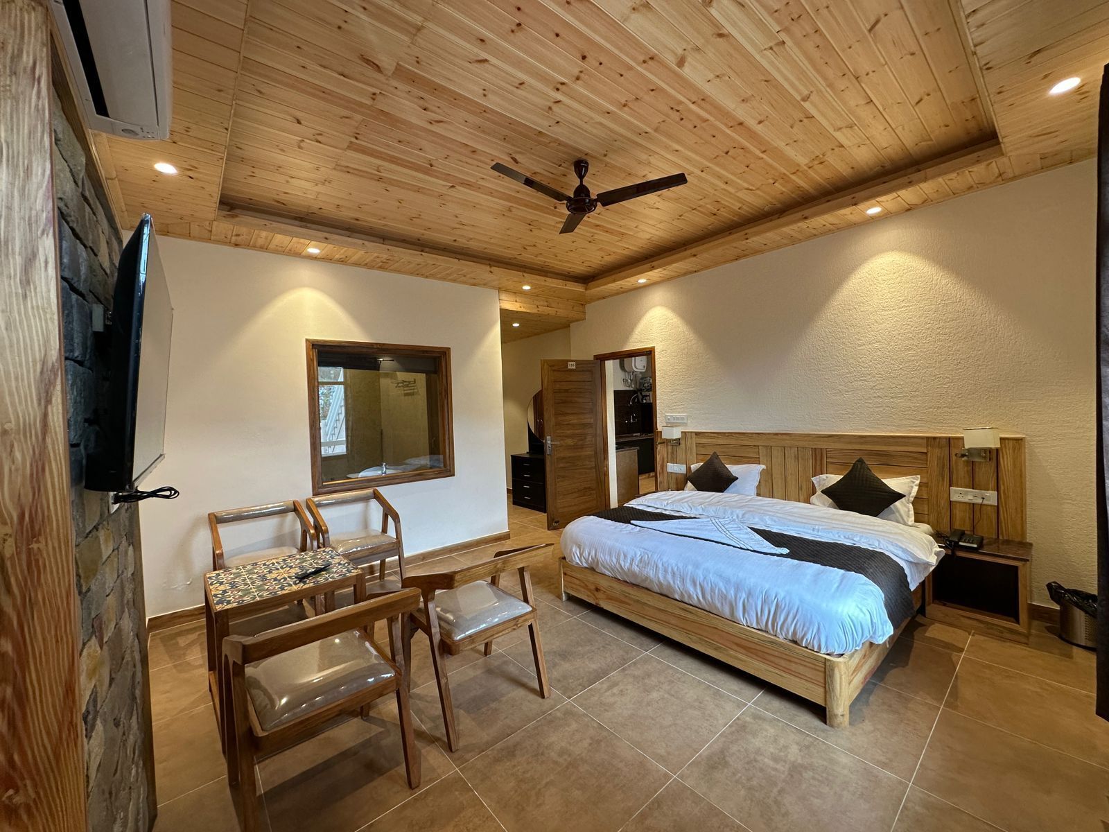 A bedroom with a wooden ceiling, a bed with white bedding, a wooden desk with a chair, and a television mounted on the wall.