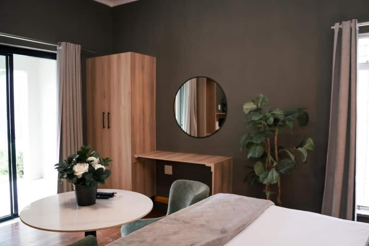 A modern hotel room with a bed, a round table, a desk, a mirror, and a potted plant.