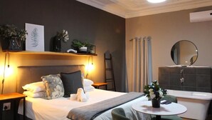 A hotel room with a bed, bedside table, lamp, wall-mounted shelf with plants, a round table with a flower vase, and a bathroom with a bathtub and mirror. - Stay Local Potchefstroom (Potcherfstroom)