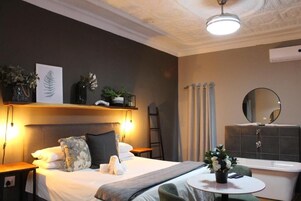 A hotel room with a bed, bedside table, lamp, wall-mounted shelf with plants, a round table with a flower vase, and a bathroom with a bathtub and mirror.