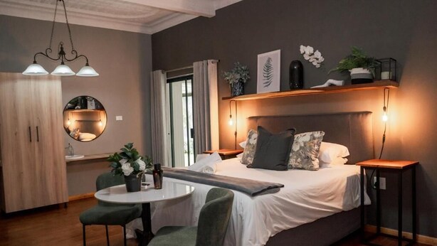 A modern bedroom with a bed, bedside tables, a round mirror, a small table with chairs, and a view of the outside. - Stay Local Potchefstroom (Potcherfstroom)