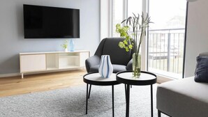 A living room with a flat-screen TV, a grey sofa, a small table with a vase, and a white console table. - To port (Sankt Peter-Ording)
