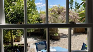 A window with a view of a garden, outdoor seating, and a building in the background. - Laundry Cottage: a little piece of heaven in North Cornwall! (Bodmin)