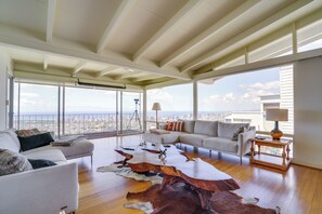 A spacious living room with a high ceiling, large windows offering a city view, a leather coffee table, and a sofa.