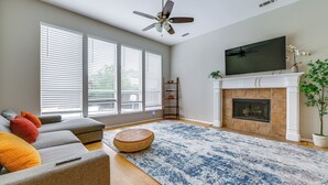 A living room with a sofa, a round wooden ottoman, a fireplace, and a television mounted above it. - 4 Mi to Toyota Stadium! Family Haven w/ Pool Oasis (Frisco)