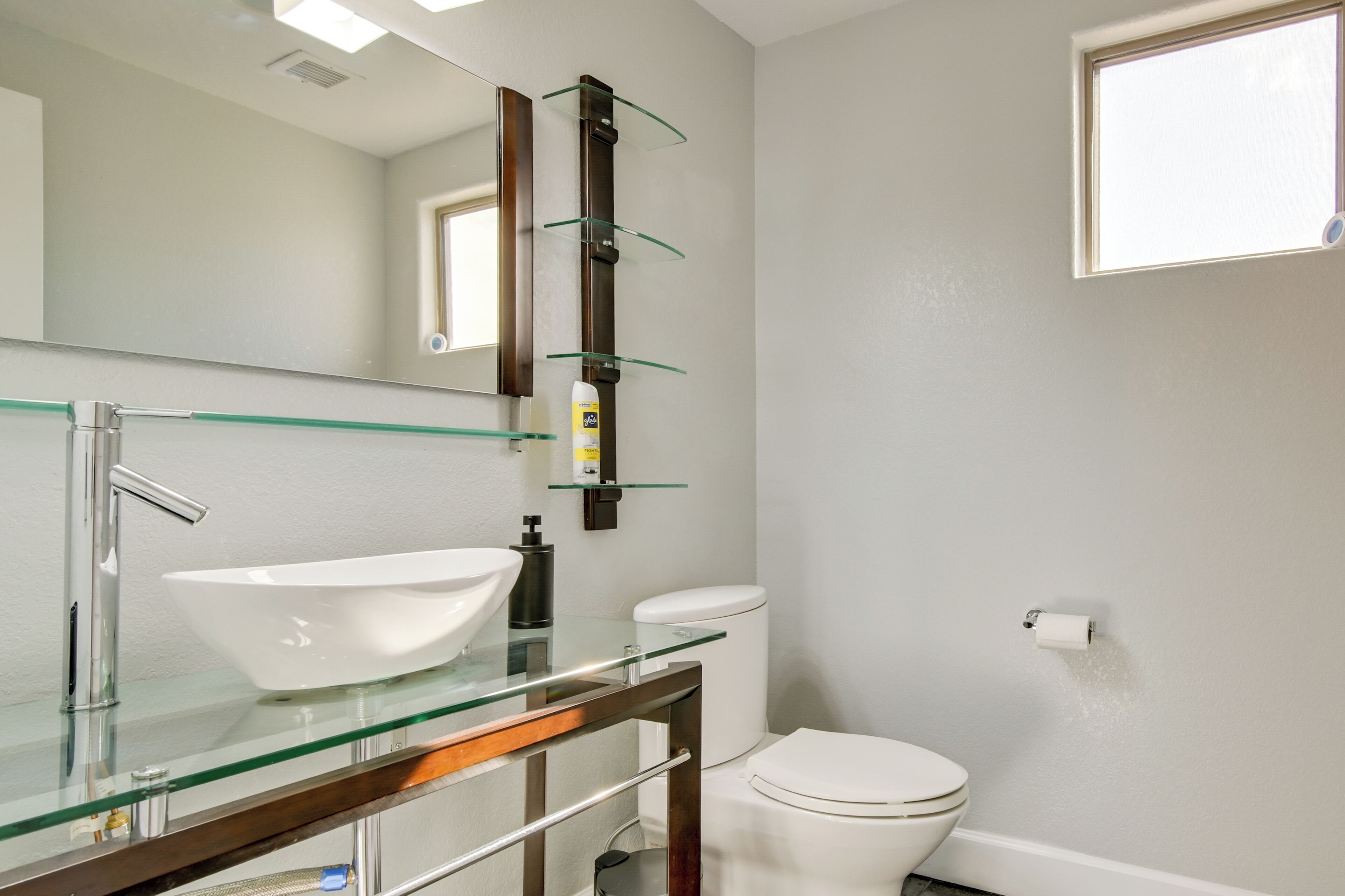 A bathroom with a glass sink, a mirror, a toilet, and a shelf with toiletries.