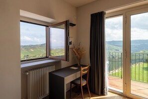 Ein Zimmer mit Schreibtisch, Stuhl, Heizkörper, einem Fenster mit Blick ins Grüne und einem Balkon mit Geländer.