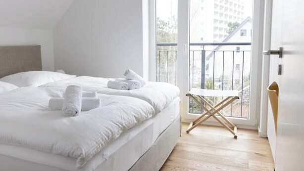 A bedroom with a bed, a window with a view of buildings, and a balcony. - About Strandroggen (Sankt Peter-Ording)