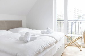 A hotel room with a bed, pillows, a chair, and a view of a building through a window. - About Strandroggen (Sankt Peter-Ording)