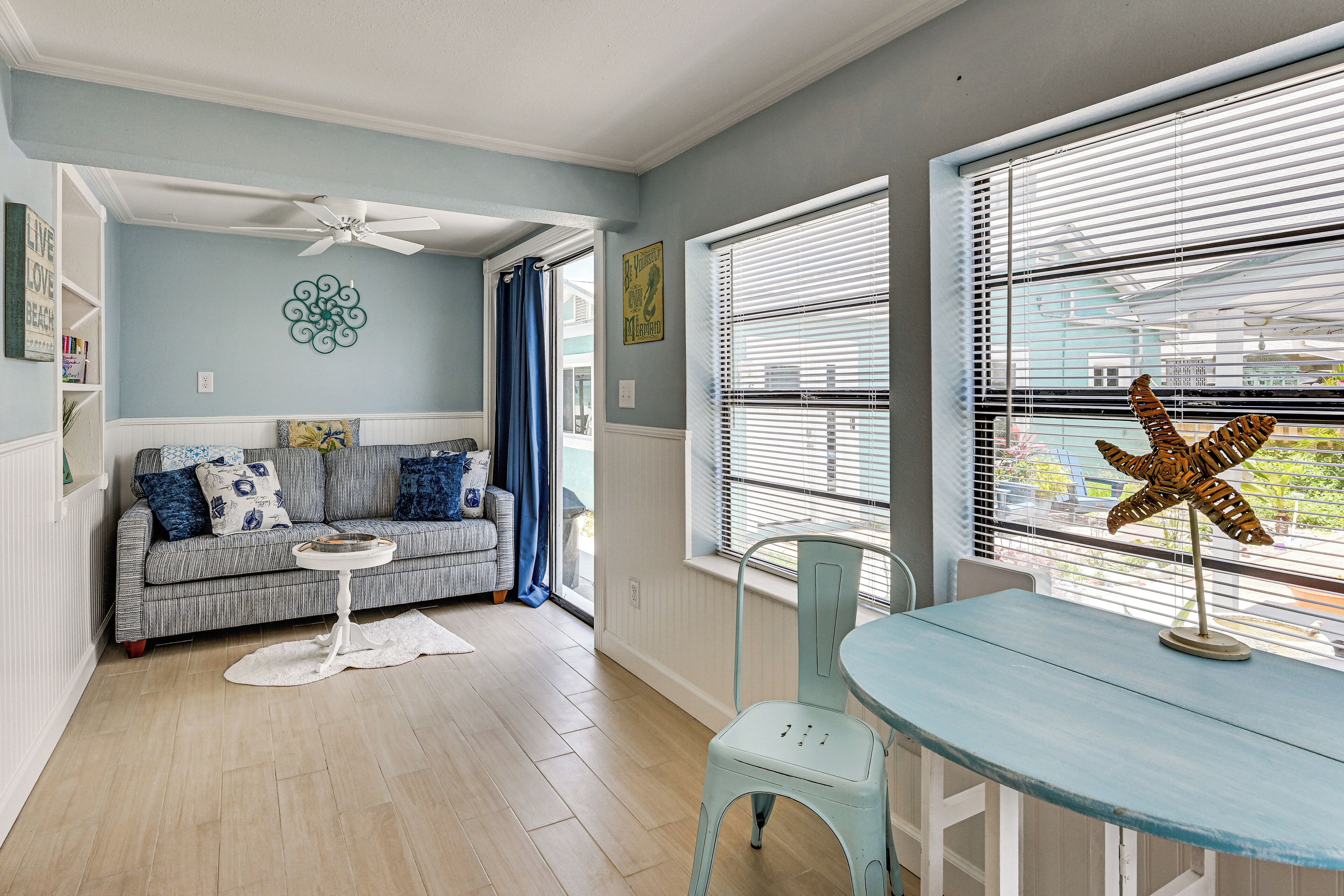 A room with a sofa, a small table, a ceiling fan, and a window with blinds.
