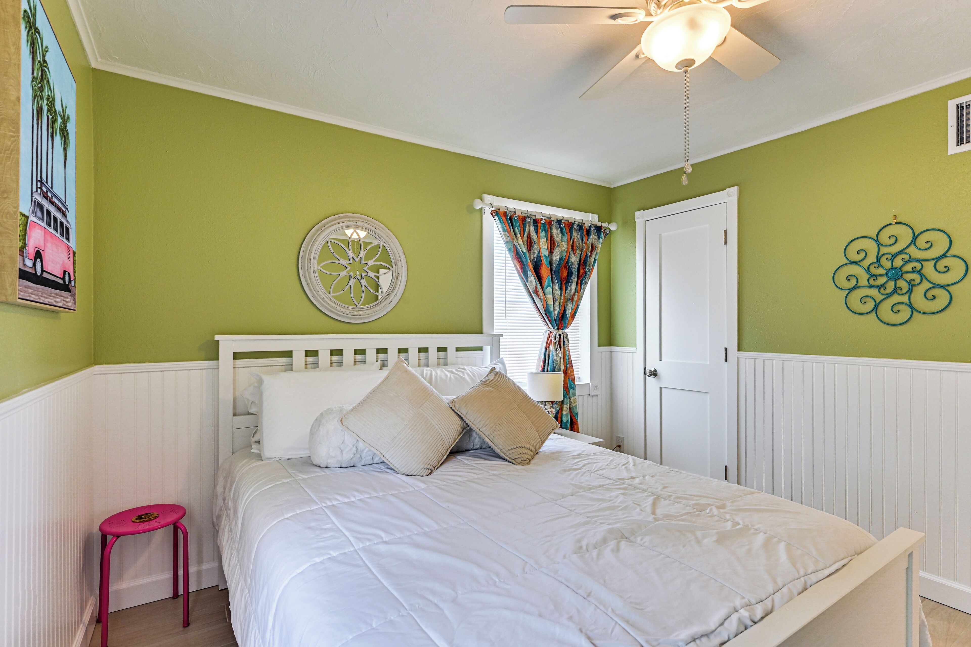 A bedroom with a bed, a ceiling fan, a window with curtains, a door, and a wall decoration.