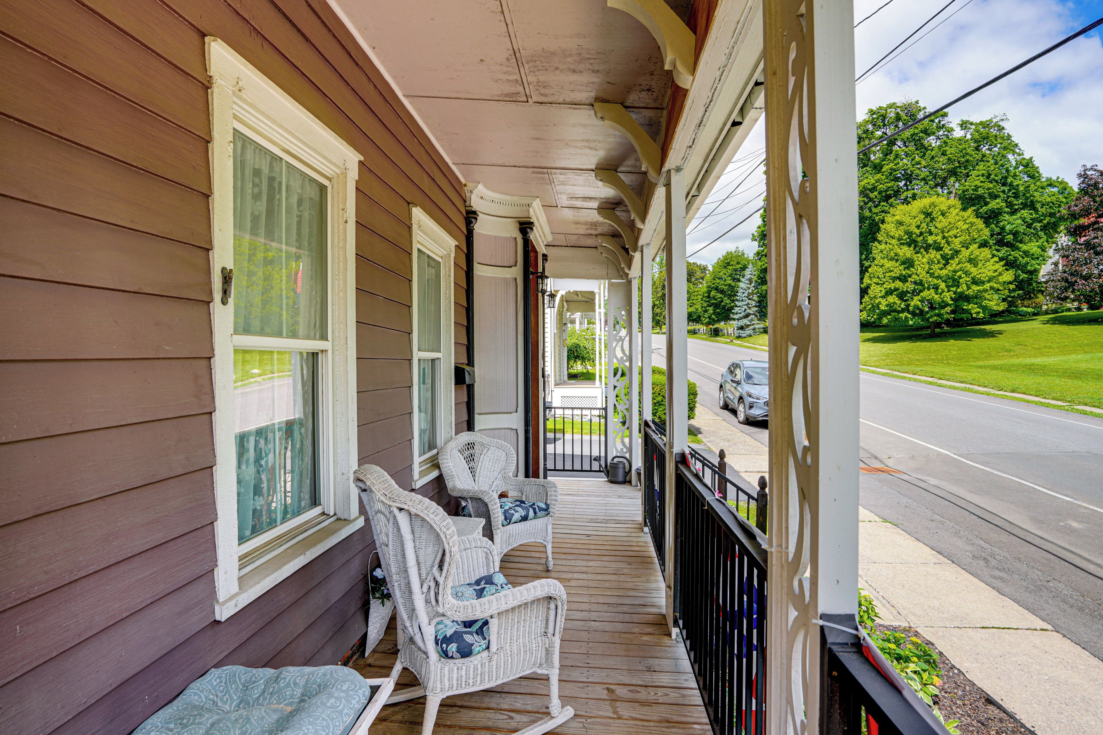 A porch with two rocking chairs, a covered walkway, and a view of a street with cars and greenery.