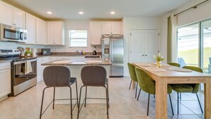 A modern kitchen with white cabinets, a stainless steel refrigerator, and a dining area with a wooden table and green chairs.