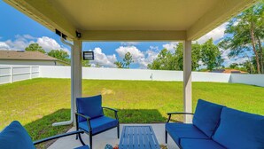 A covered patio with a table and two blue chairs, a white fence, and a grassy yard.