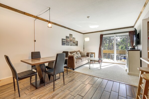 Un salon comprenant une table à manger, des chaises, un canapé, une table basse et un téléviseur. Il y a un balcon avec vue sur les arbres.
