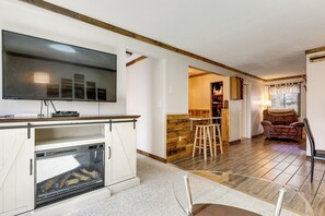A living room with a TV, fireplace, wooden floor, and a dining area with stools. - Walk to Weirs Beach! Family Escape w/ Balcony (Laconia)