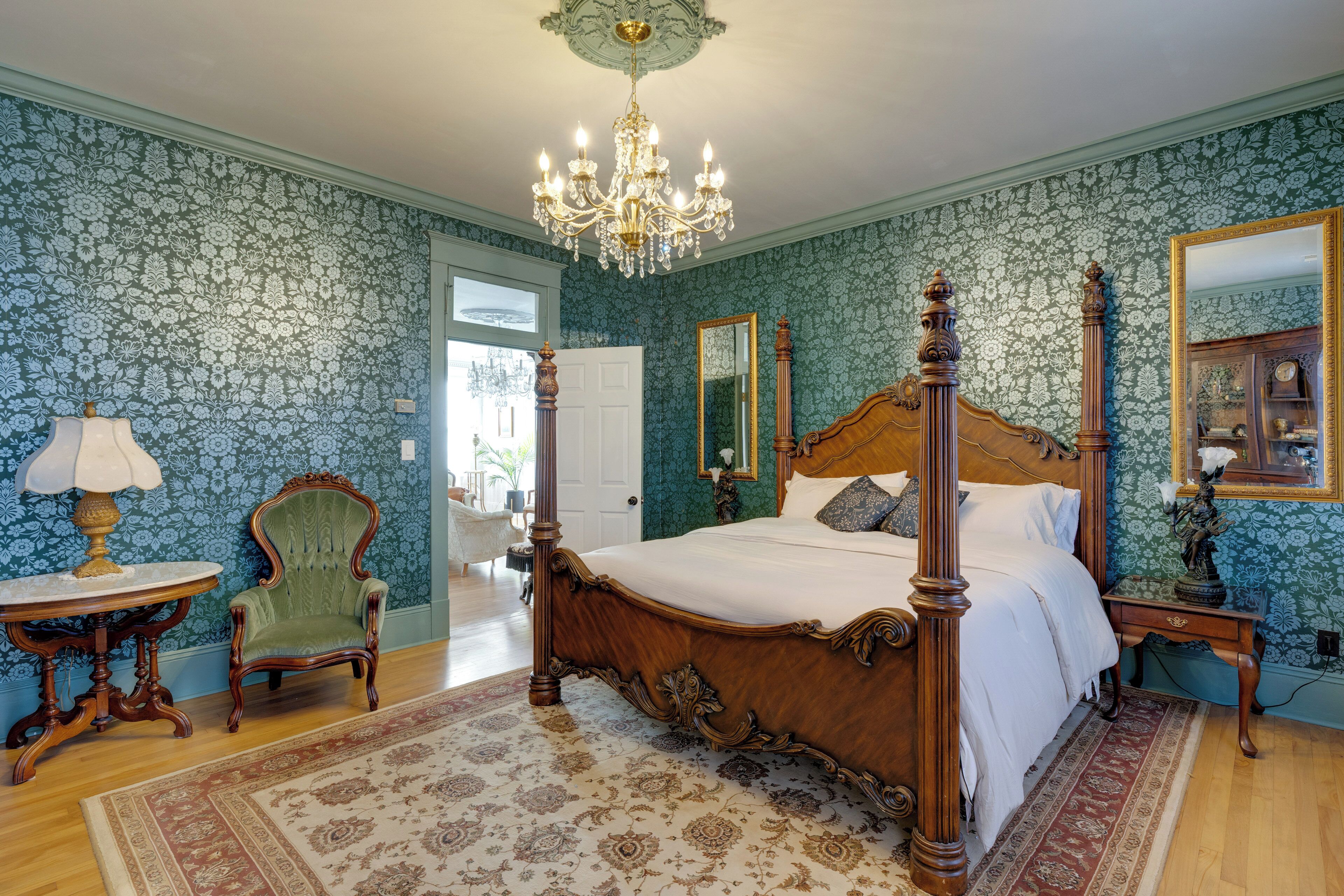 A bedroom with a four-poster bed, a chandelier, a green armchair, and a mirror.