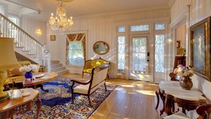 A grand room with a staircase, a chandelier, a yellow armchair, and a large window with decorative glass.