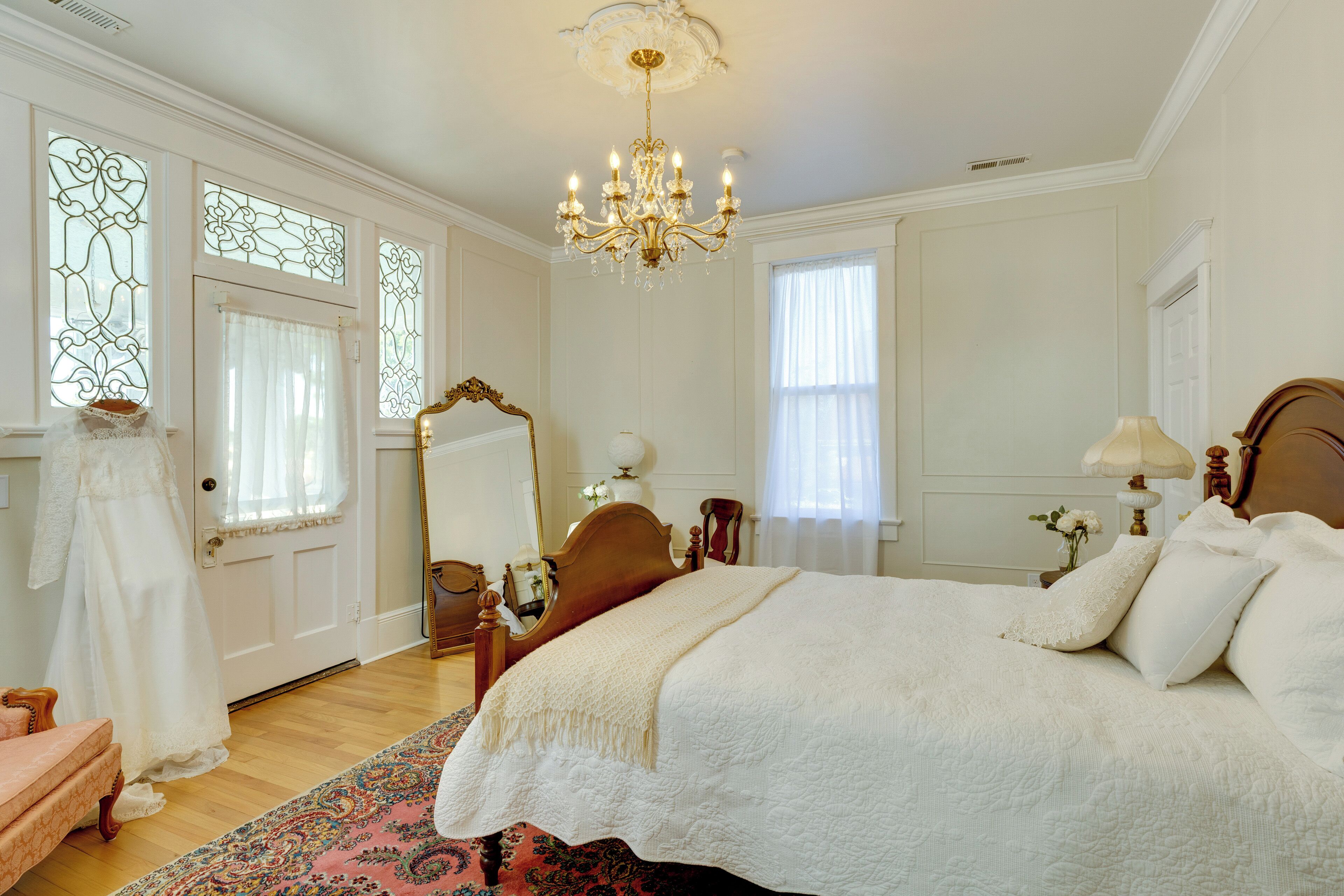 A bedroom with a large bed, a chandelier, a mirror, and a dress on the floor.
