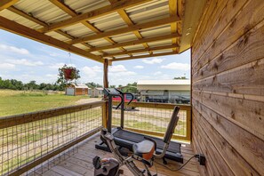 Een overdekt buitengebied met fitnessapparatuur, een houten terras en uitzicht op een grasveld en voertuigen op de achtergrond.