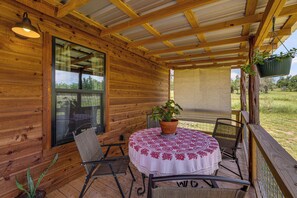 A covered patio with a round table, chairs, a hanging plant, and a window. - Romantic Wine Country Cabin Near Fredericksburg (Harper)