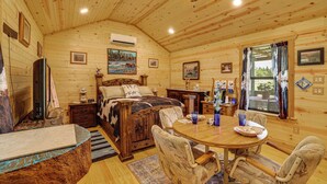 Interior de una cabaña de madera con una cama, mesa de comedor, sillas y un ventilador de techo.