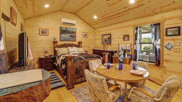 A wooden cabin interior with a bed, dining table, chairs, and a ceiling fan. - Romantic Wine Country Cabin Near Fredericksburg (Harper)