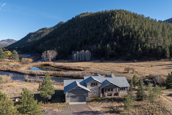 A two-story stone house with a garage, surrounded by a natural landscape with a river and forested hills.