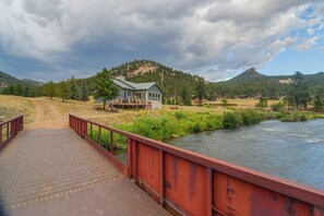 Eine rustikale Brücke führt zu einem Haus mit Stein Fundament, umgeben von einem Fluss und einer Berglandschaft.