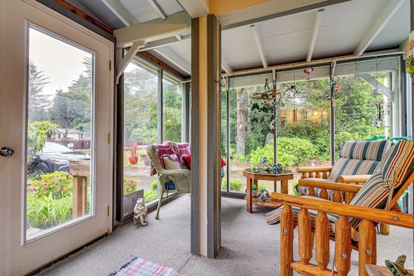 A room with a large sliding glass door, a wicker chair, a wooden bench with striped cushions, and a small table with decorative items. - 1 Mi to Beach! Lush Florence Retreat w/ Sunroom (Florence)