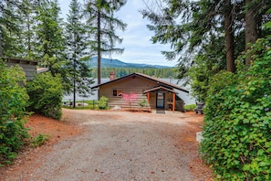 A cabin with a gravel driveway, surrounded by dense forest and mountains in the background.