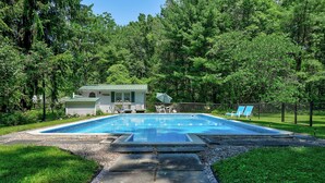 A swimming pool with a stone pathway leading to it, surrounded by lush greenery and a small cabin.