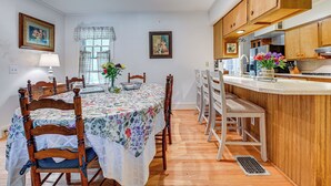 A dining area with a table set for four, wooden chairs, a kitchen with wooden cabinets, and a window with curtains.