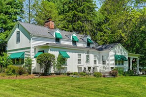A white two-story house with green awnings, a front porch with a chair, and a brick chimney.